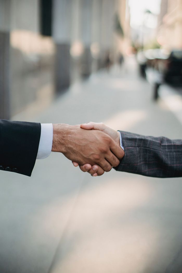 who-we-are Two businessmen shaking hands outside, symbolizing cooperation and agreement in a city setting.